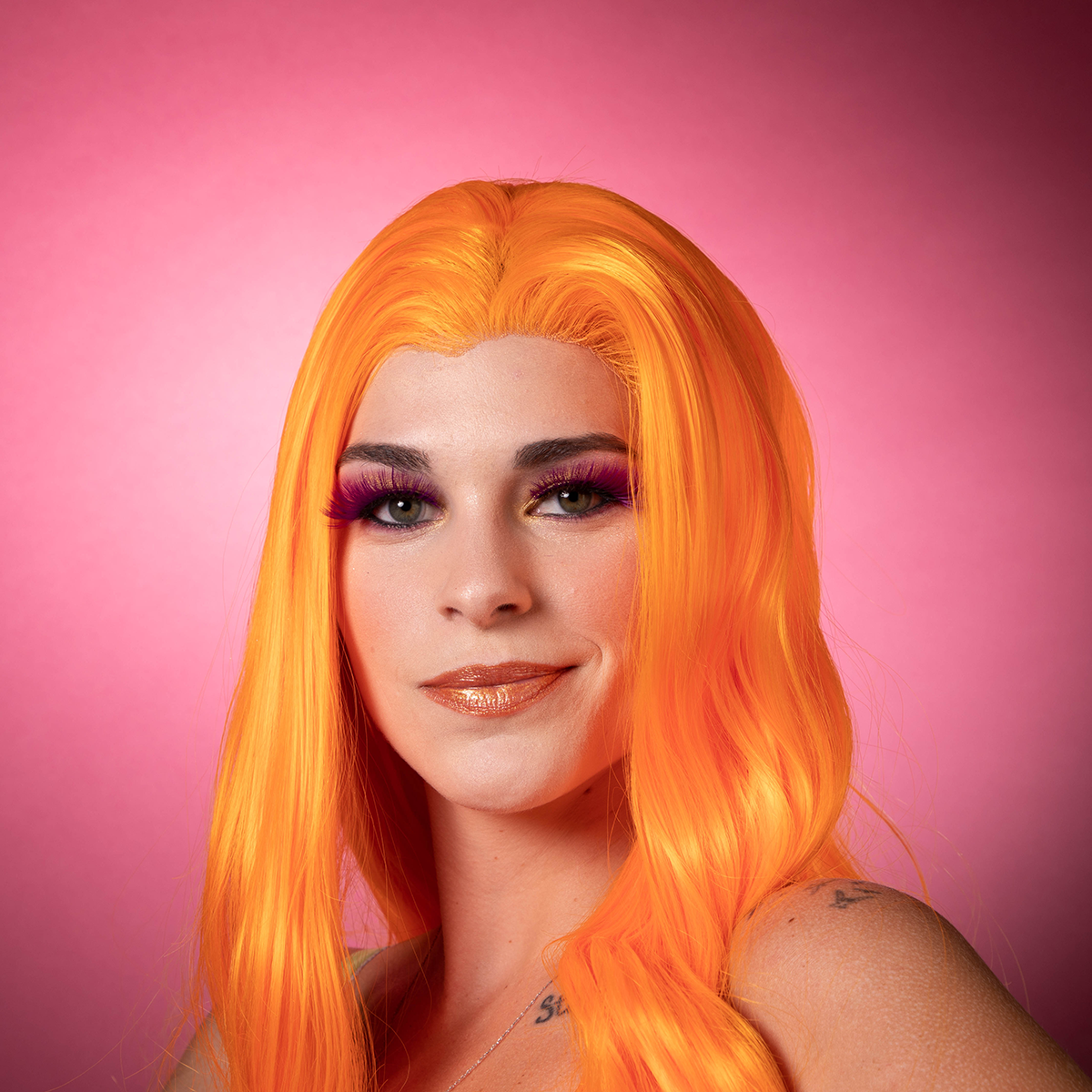 Person with bright orange hair and colorful makeup against a pink background