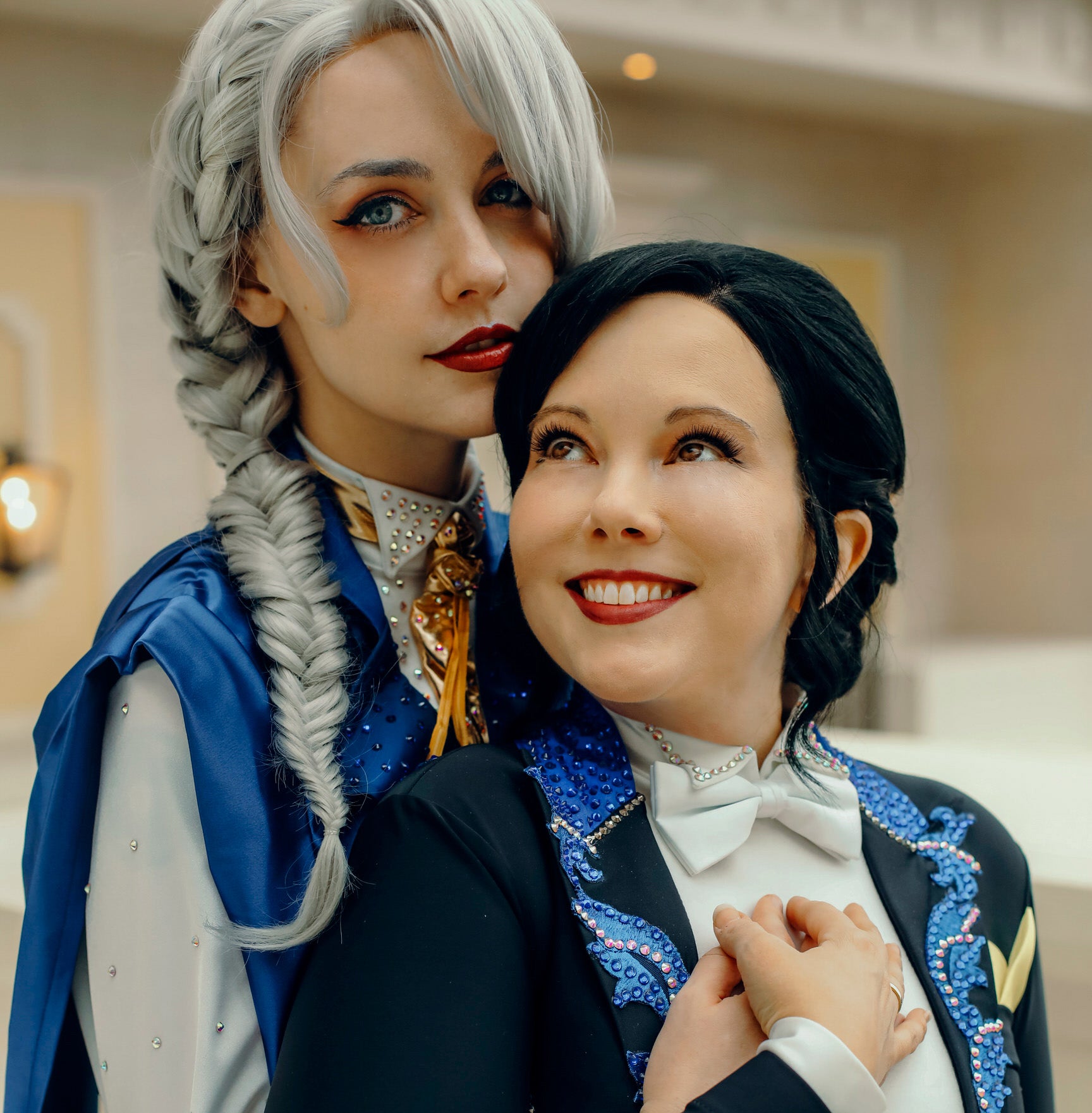 Two people in elaborate costumes with decorative elements, one with gray hair and blue outfit, the other with black hair and dark outfit.